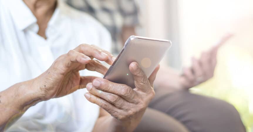 Closeup of elderly woman's hands using a mobile phone with elderly man's hands and phone blurred in the background, illustrating the blog topic of Top Free Apps for Seniors on Medicare